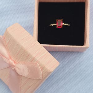 Op de afbeelding: Een gouden ring met een rechthoekige rode edelsteen, gepresenteerd in een open roze geschenkdoos met een zwart interieur. De ring is versierd met kleine, heldere stenen. De doos is vastgebonden met een roze lint.