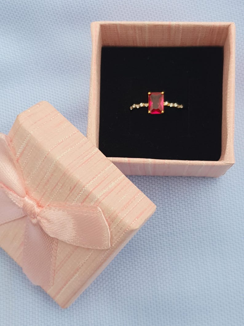 Op de afbeelding: Een gouden ring met een rechthoekige rode edelsteen, gepresenteerd in een open roze geschenkdoos met een zwart interieur. De ring is versierd met kleine, heldere stenen. De doos is vastgebonden met een roze lint.
