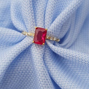 Op de afbeelding: Een gouden ring met een grote, rechthoekige, rode edelsteen. De band is versierd met kleine, heldere edelstenen. De ring is geplaatst tegen een lichtblauwe stoffen achtergrond, wat een contrast van kleuren en texturen cre&euml;ert.