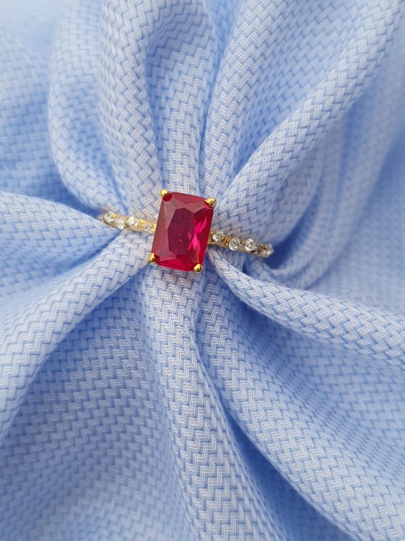 Op de afbeelding: Een gouden ring met een grote, rechthoekige, rode edelsteen. De band is versierd met kleine, heldere edelstenen. De ring is geplaatst tegen een lichtblauwe stoffen achtergrond, wat een contrast van kleuren en texturen cre&euml;ert.