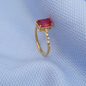 Op de afbeelding: Een gouden ring met een rechthoekige, rode edelsteen en kleine, heldere accentstenen. De ring is geplaatst tegen een lichtblauwe stoffen achtergrond.