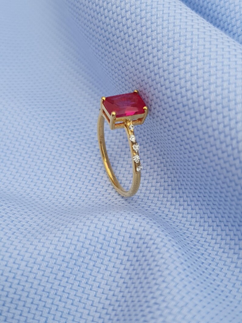 Op de afbeelding: Een gouden ring met een rechthoekige, rode edelsteen en kleine, heldere accentstenen. De ring is geplaatst tegen een lichtblauwe stoffen achtergrond.