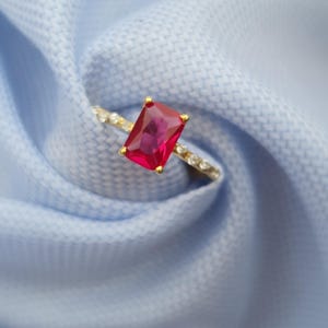 Op de afbeelding: Een gouden ring met een grote, rechthoekige, robijnrode edelsteen. De band van de ring is versierd met kleine, heldere edelstenen. De ring is geplaatst tegen een lichtblauwe stoffen achtergrond.