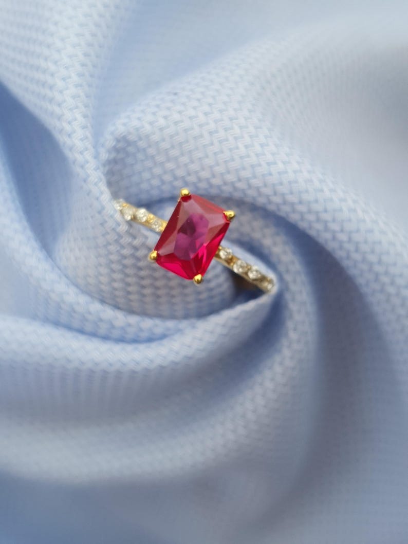Op de afbeelding: Een gouden ring met een grote, rechthoekige, robijnrode edelsteen. De band van de ring is versierd met kleine, heldere edelstenen. De ring is geplaatst tegen een lichtblauwe stoffen achtergrond.