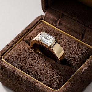 May include: A gold ring featuring a single emerald-cut clear gemstone set in a polished yellow gold band. The ring is displayed inside a dark brown velvet-lined jewelry box with gold trim along the inner edge of the lid and base.