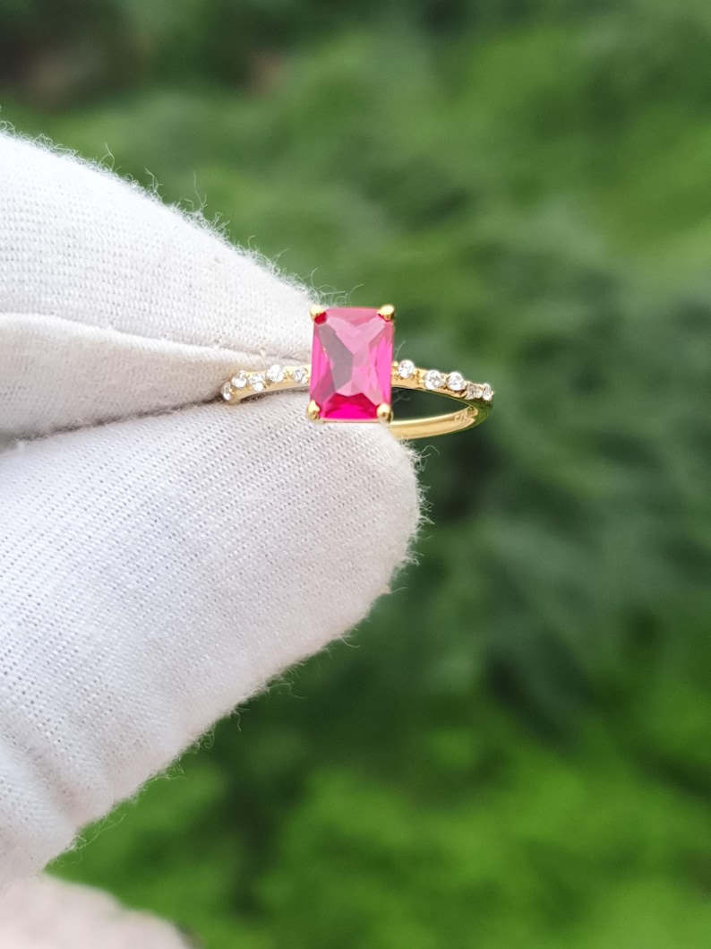 Op de afbeelding: Een gouden ring met een grote, rechthoekige, helder roze edelsteen. De band is gesplitst, met kleine, heldere stenen langs de splitsing. De ring wordt gepresenteerd tegen een vage groene achtergrond.
