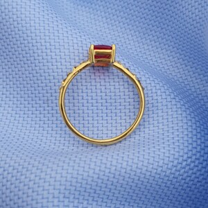 Op de afbeelding: Een gouden ring met een rechthoekige, rode edelsteen. De band is versierd met kleine, sprankelende accenten. De ring is geplaatst tegen een lichtblauwe stoffen achtergrond, wat een contrast cre&euml;ert.
