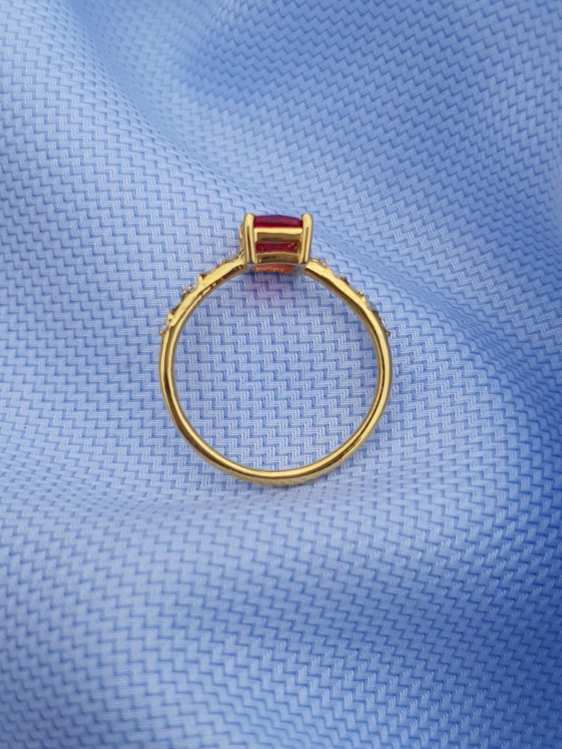 Op de afbeelding: Een gouden ring met een rechthoekige, rode edelsteen. De band is versierd met kleine, sprankelende accenten. De ring is geplaatst tegen een lichtblauwe stoffen achtergrond, wat een contrast cre&euml;ert.