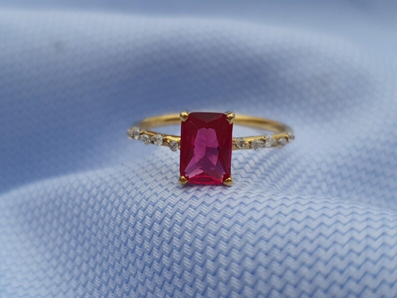 Op de afbeelding: Een gouden ring met een grote, rechthoekige, robijnkleurige edelsteen als middelpunt. De band is ook versierd met kleinere, heldere edelstenen. De ring staat tegen een lichtblauwe stoffen achtergrond.