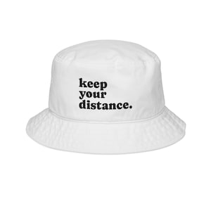 May include: White bucket hat with the text "keep your distance" in black, centered on the front. The hat has a wide brim and a rounded crown. This hat is a casual accessory.