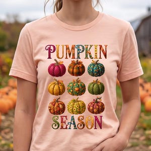 May include: A peach-colored short-sleeve t-shirt featuring the text PUMPKIN SEASON in colorful, patterned lettering. The design includes a grid of nine illustrated pumpkins, each displaying unique textures, colors, and patterns like plaid and floral.