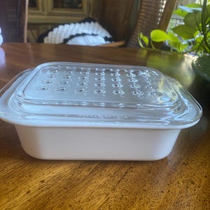 May include: A white rectangular Pyrex baking dish with a clear glass lid. The lid has a series of small, evenly spaced holes. The dish sits on a wooden surface.