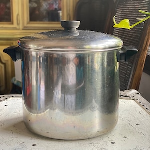 May include: A large, stainless steel cooking pot with a lid and black handles. The pot is cylindrical and reflects the surrounding environment. The lid has a black knob. The pot sits on a white, distressed surface.