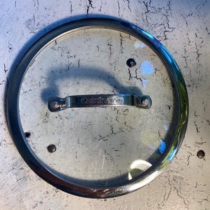 May include: A clear glass lid with a silver metal rim and handle. The lid has the word "Cuisinart" printed on the handle.