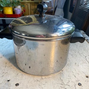 May include: A silver stainless steel pot with black handles and a black knob on the lid.