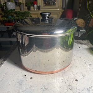 May include: A stainless steel pot with a copper bottom and a black handle. The pot has a shiny, reflective surface and a lid with a black knob. The pot is sitting on a white, distressed surface.