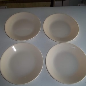 May include: Four off-white, round plates are arranged in a square formation. The plates have a slightly curved rim and a smooth, glossy surface. The plates are a neutral color, suitable for various dining settings.
