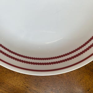 May include: White ceramic plate with a maroon dotted border design. The plate has a simple, elegant aesthetic, suitable for everyday use or special occasions. The maroon border adds a touch of color and visual interest to the otherwise plain white surface.