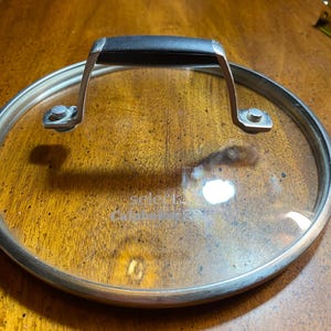 May include: A clear glass pot lid with a stainless steel rim and a black handle. The lid has a silver metal handle attached with rivets. The text "select" and "Calphalon" is visible on the glass.