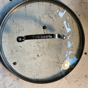 May include: A clear glass lid with a silver metal rim and handle. The handle has the word "Cuisinart" printed on it.