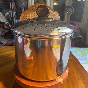 May include: A stainless steel pot with a perforated lid and black handles. The pot sits on a round wooden trivet. The pot is reflective, showing the surrounding environment. A dark brown knob is on top of the lid.