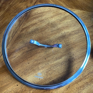 May include: A clear glass lid with a silver-colored metal rim and handle. The lid is circular and designed for cookware. The handle is a curved shape with two rounded ends. The lid is sitting on a wooden surface.