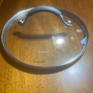 May include: A clear glass Cuisinart lid with a stainless steel rim and a black handle. The lid is round and designed for cookware. The handle is curved for easy gripping. The Cuisinart logo is visible on the glass.