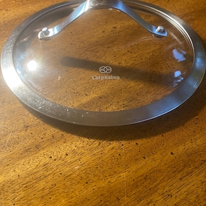 May include: A clear glass Calphalon brand pot lid with a stainless steel rim and handle. The lid is round and has a silver handle attached with two rivets. The Calphalon logo is visible on the glass.