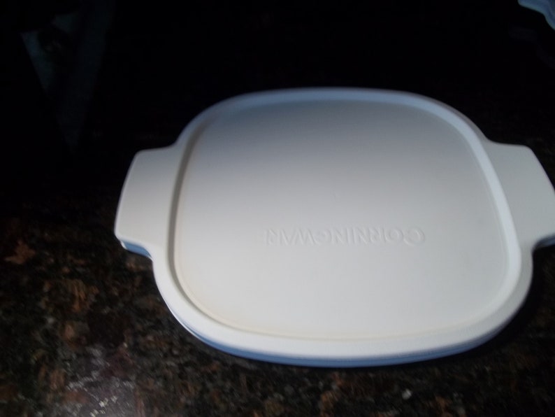 May include: A white CorningWare lid with a rectangular shape and rounded corners. The lid has two handles on opposite sides and the brand name is embossed on the surface. The lid is designed for cookware.