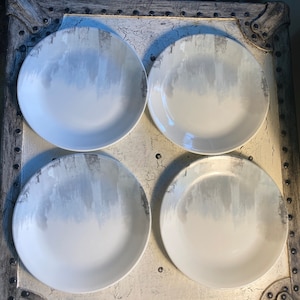 May include: Four white ceramic dinner plates with a gray ombre design. The plates are arranged in a square pattern on a distressed white surface.