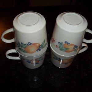 Puede incluir: Juego de cuatro tazas de cer&aacute;mica blanca con un dise&ntilde;o de frutas y flores. Las tazas est&aacute;n apiladas en pares.