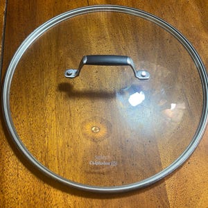 May include: A clear glass Calphalon Select pot lid with a stainless steel rim and a black handle. The lid is circular and sits on a wooden surface.