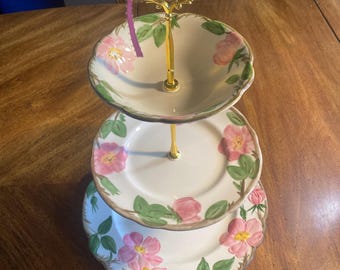 May include: A three-tiered serving stand featuring white ceramic plates with scalloped edges, decorated with pink floral patterns and green leaves. The stand has a central gold-tone metal rod and a decorative handle with a purple ribbon tied to it.