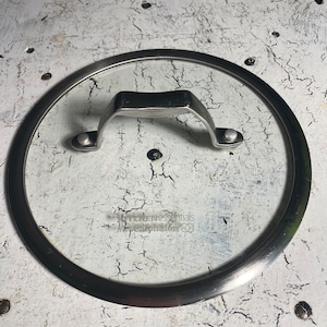 May include: A silver-rimmed glass lid with a metal handle. The lid is sitting on a white surface with a cracked texture.