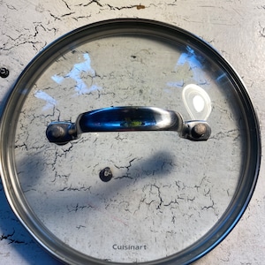 May include: A silver-colored glass lid with a handle and the word "Cuisinart" printed on the bottom.