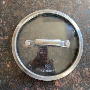 May include: A round, clear glass Calphalon brand pot lid with a stainless steel rim and handle. The lid is designed to fit a pot and has a silver handle attached with two rivets.