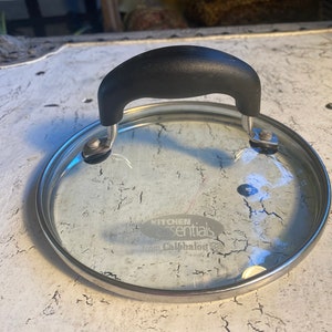 May include: A clear glass lid with a silver rim and a black handle. The lid has a sticker that says "Kitchen Essentials from Calphalon".