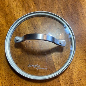 May include: A clear glass lid with a stainless steel rim and handle. The lid has the text "Simply Calphalon" printed on it. The lid is designed for cookware.