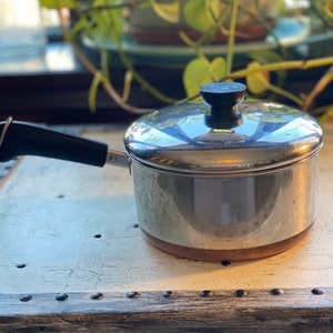 May include: A silver saucepan with a copper bottom and a black handle. The saucepan has a lid with a black knob.