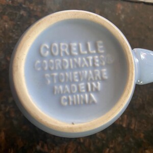 May include: A light blue Corelle mug, viewed from the bottom, with the words "CORELLE COORDINATES STONEWARE MADE IN CHINA" embossed in white. The mug has a light beige rim.