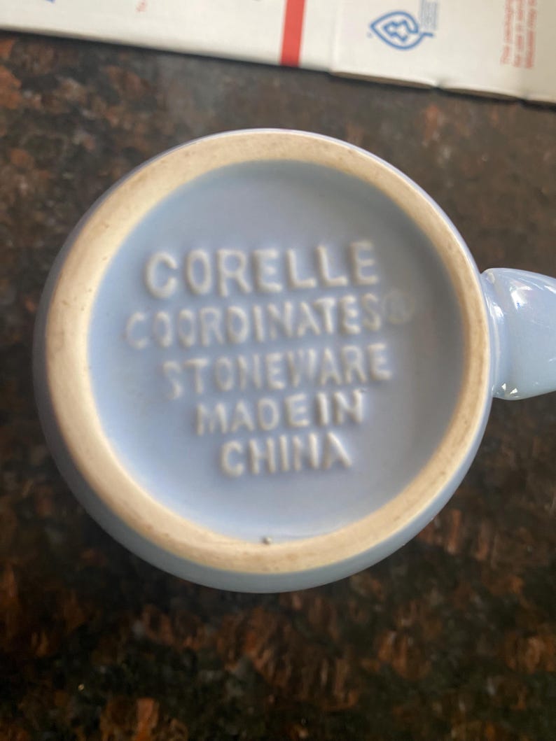 May include: A light blue Corelle mug, viewed from the bottom, with the words "CORELLE COORDINATES STONEWARE MADE IN CHINA" embossed in white. The mug has a light beige rim.