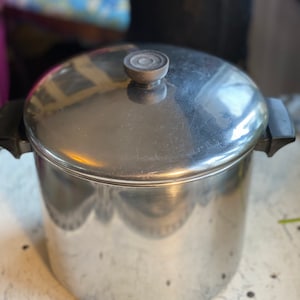May include: A stainless steel cooking pot with a lid. The pot has two black handles and a round knob on the lid. The pot is sitting on a white surface.