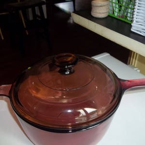 May include: A reddish-brown glass saucepan with a matching lid and handle. The lid has a dark knob. The pan is sitting on a white surface. The handle extends to the right.