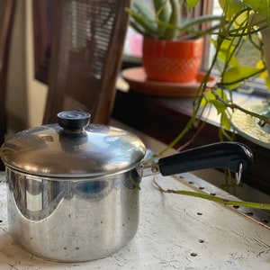 May include: A silver saucepan with a black handle and lid. The saucepan is sitting on a white table with a distressed finish.