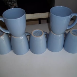 May include: Six light blue ceramic mugs. Two mugs are upright, with handles visible, stacked on top of four inverted mugs. The mugs have a smooth, glossy finish and a simple, classic design. The mugs are arranged on a white surface.