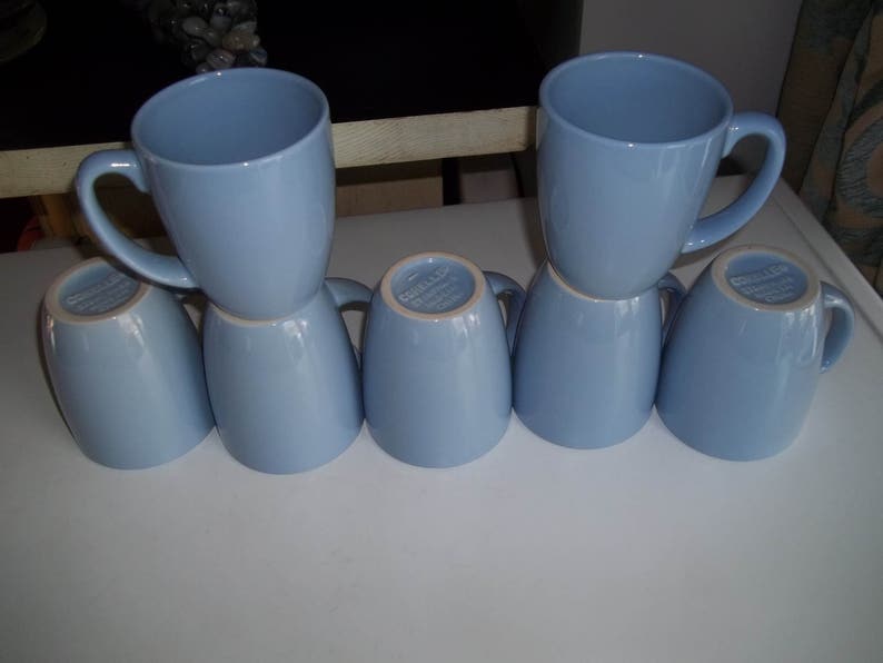 May include: Six light blue ceramic mugs. Two mugs are upright, with handles visible, stacked on top of four inverted mugs. The mugs have a smooth, glossy finish and a simple, classic design. The mugs are arranged on a white surface.