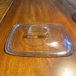May include: A rectangular clear glass lid featuring rounded corners and a centered, integrated glass handle. The lid has a raised outer rim designed to fit securely onto a corresponding container, providing a transparent cover for kitchen storage.