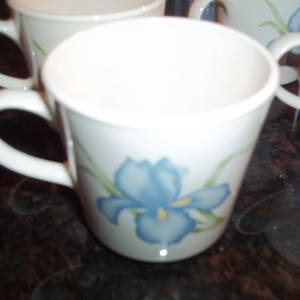 May include: A white ceramic mug with a blue and green floral design. The mug has a handle and is decorated with a blue iris flower.