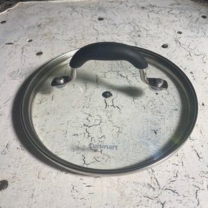 May include: A clear glass lid with a black handle and a silver rim. The lid has the word "Cuisinart" printed on it.