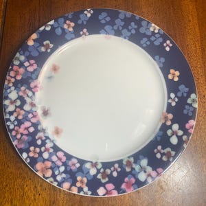 May include: A round white plate with a dark blue floral border. The border features a pattern of small, colorful flowers in shades of pink, blue, and peach. The plate is set against a wooden surface.